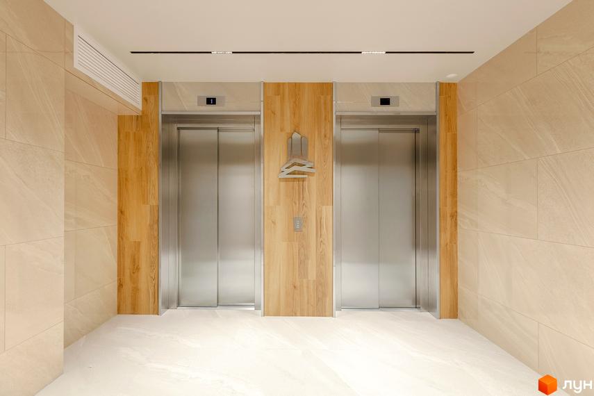 Два ліфти в сучасному холі ЖК Obolon Plaza. Оздоблення стін мармуровою плиткою та дерев'яними панелями. Біла підлога.