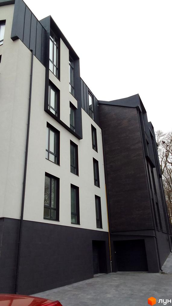 Зовнішній вигляд новобудови по вул. Петрушевича, 8 з білою та чорною цегляною обробкою та великими вікнами. Територія навколо будинку вимощена плиткою.