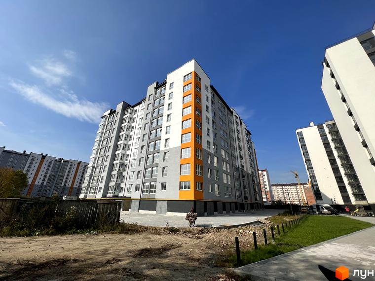 Новобудова ЖК Галицький 2 з сіро-білими фасадами та помаранчевими акцентами. Навколо новобудови облаштована прибудинкова територія з зеленими насадженнями та доріжками.