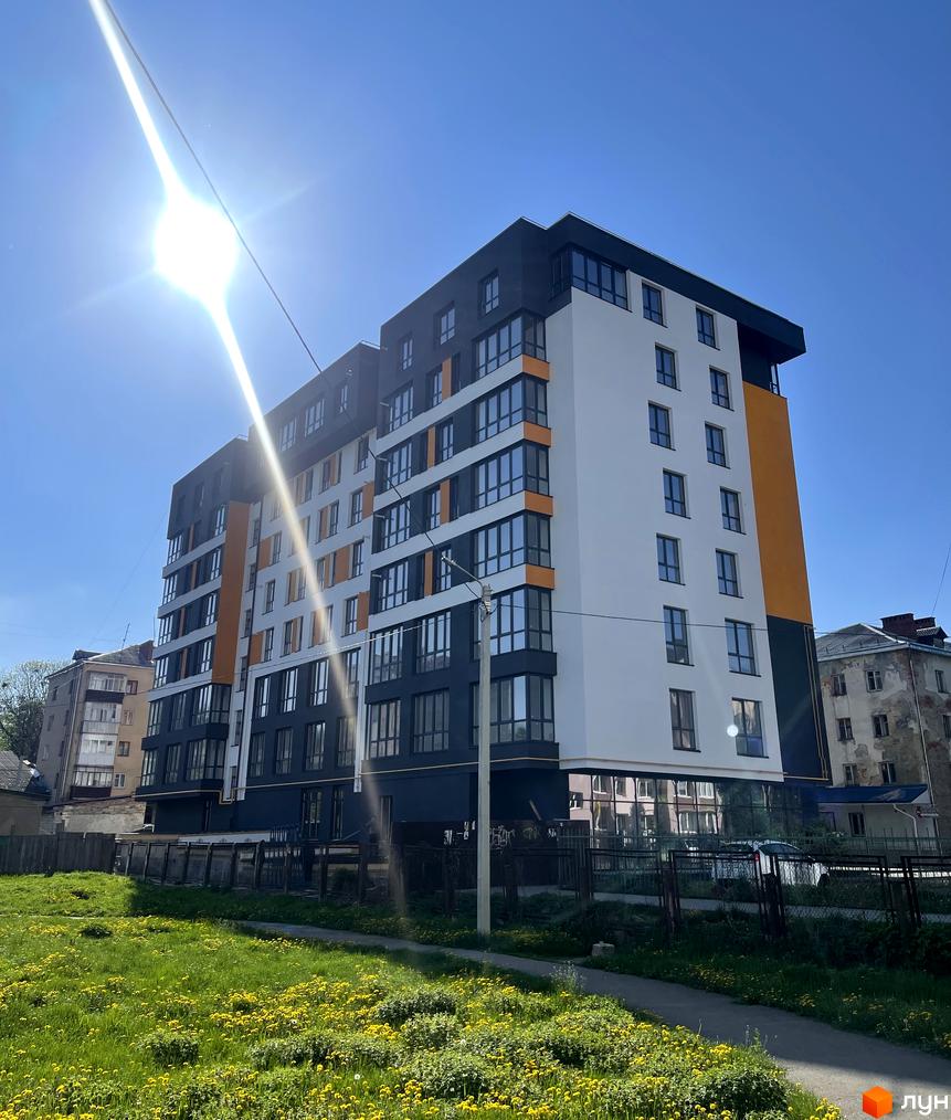 Житловий комплекс Квартал Галичанка - це сучасна новобудова з біло-темно-помаранчевим фасадом та великими вікнами. Перед будинком розташований зелений двір з квітами та парканом.