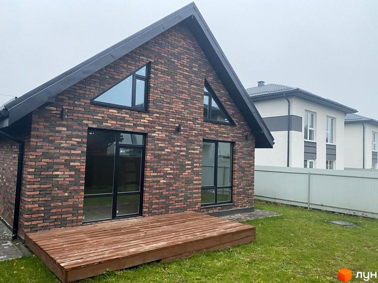 Котеджне містечко Одноповерховий Київ - Віта-Поштова. Будинок з червоної цегли з великими вікнами та дерев'яною терасою на тлі зеленого газону та сусідніх будинків.