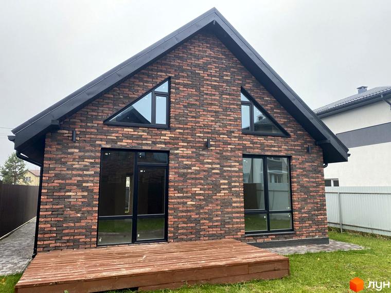 Котеджне містечко КМ Одноповерховий Київ - Віта-Поштова. Фасад будинку оздоблений цеглою, з великими вікнами та дерев'яною терасою. Навколо будинку зелена галявина та доріжка з бруківки.