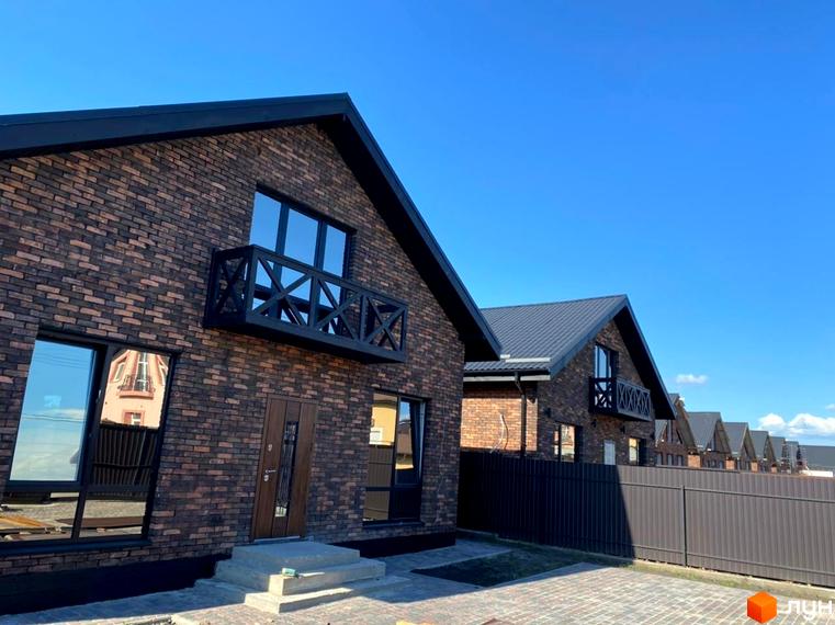 Зображення сторінки житлового комплексу КМ Південна Баварія. Цегляні будинки з чорними дахами та балконами, огороджені парканом, на тлі блакитного неба.