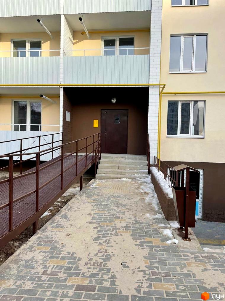 Зовнішній вигляд новобудови ЖК на Масанах з пандусом та сходами до входу. Прибудинкова територія з бруківкою та урною.