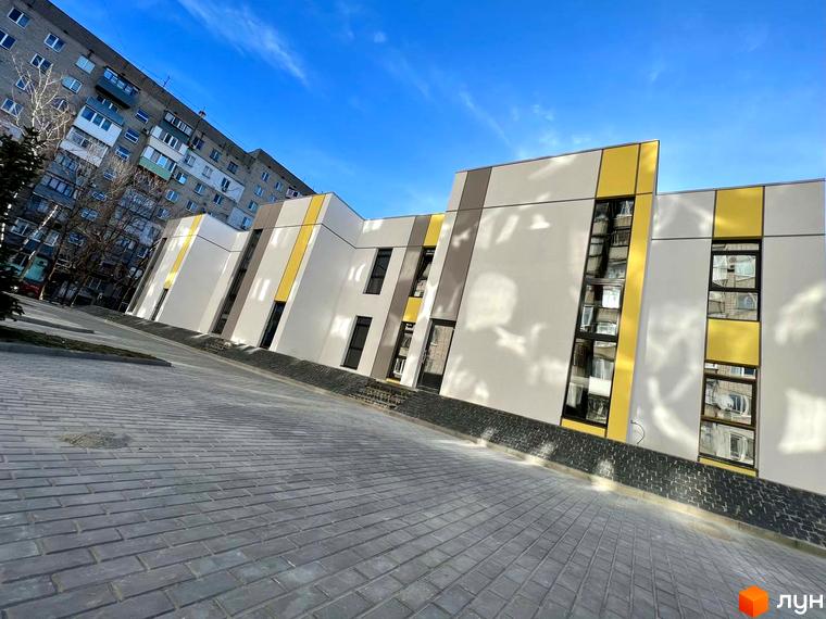 Фасад будівлі Клубний будинок ART HOUSE з білими та жовтими панелями, великими вікнами та бруківкою на прибудинковій території. Яскраве блакитне небо.