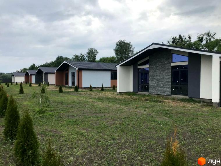 Котеджне містечко КМ Мілленіум: ряд одноповерхових будинків з темними дахами та великими вікнами, розташовані на зеленій галявині з молодими деревами.