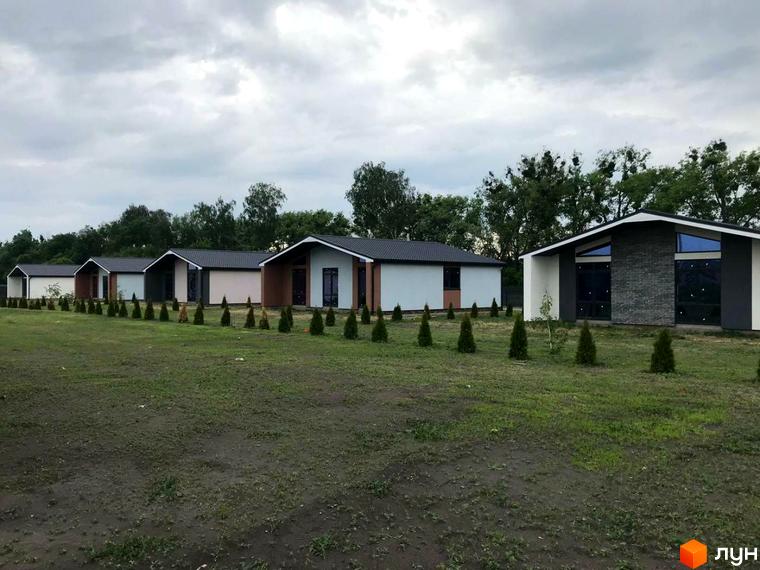 Котеджне містечко КМ Мілленіум: ряд одноповерхових будинків з темними дахами та світлими фасадами, розташованих на зеленій галявині з молодими деревами.