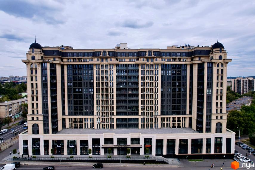 Житловий комплекс Premier Tower з бежевим та темним фасадом, що має комерційні приміщення на першому поверсі. Поруч проїжджа частина з транспортом та зелені насадження.