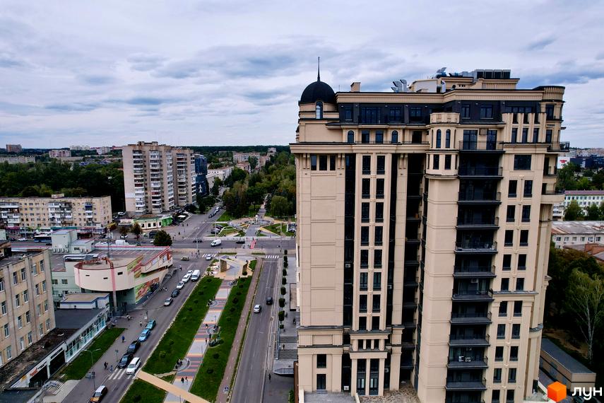 Житловий комплекс Premier Tower: багатоповерховий будинок бежевого кольору з темними вікнами та куполом. Поруч дорога, зелені зони та міська забудова.