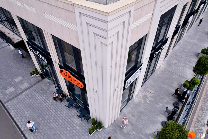 Кутовий фасад будівлі з великими вікнами та вивісками закладів. На тротуарі столики з відвідувачами та зелені насадження.