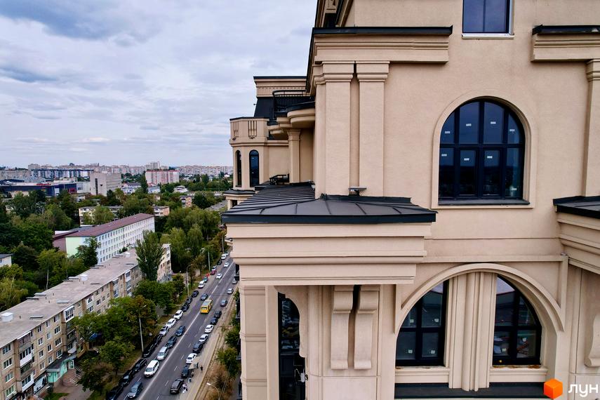 Житловий комплекс Premier Tower зі світлим фасадом, арковими вікнами та темними дахами. Поруч міська вулиця з автомобілями та багато дерев.