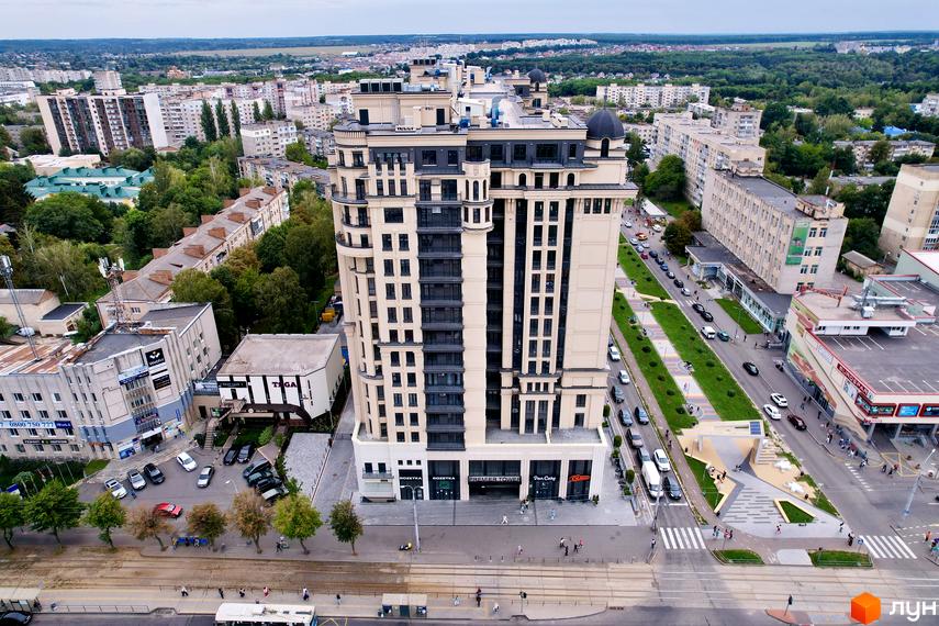 Висока будівля Житлового комплексу Premier Tower з фасадом світлого кольору та темними вікнами. Навколо міська вулиця, зелені насадження та інші будинки.