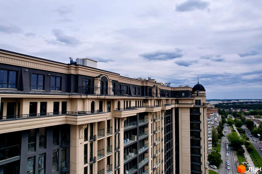 Багатоповерховий житловий комплекс Premier Tower з бежевим та темно-сірим фасадом, балконами та кутовою вежею, поруч з дорогою та зеленою зоною.