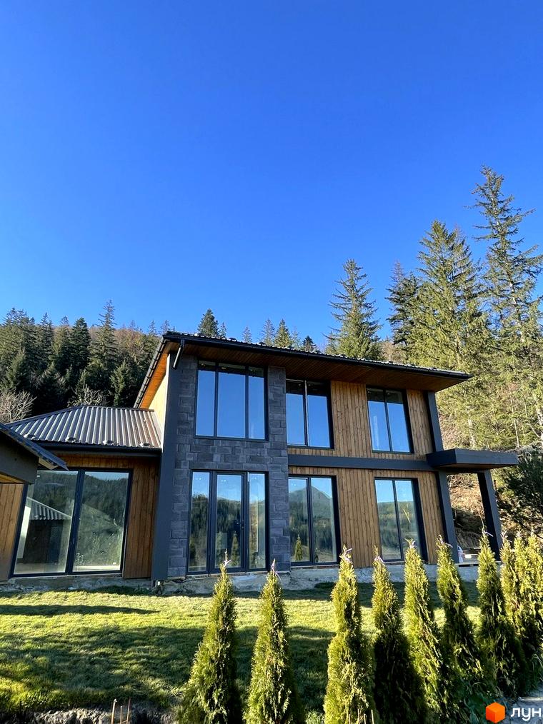 Котеджне містечко Агат Резорт & SPA: двоповерховий будинок з великими вікнами, оздоблений деревом та каменем. Перед ним зелений газон та ряд хвойних дерев на тлі лісу.