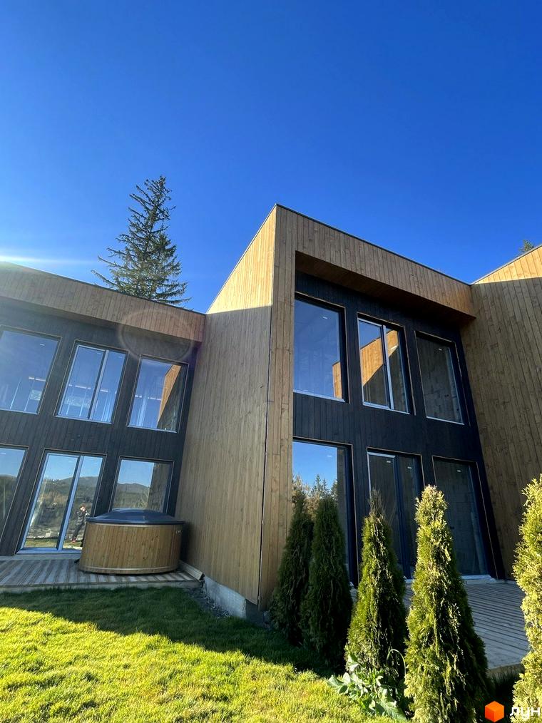 Котеджний комплекс КМ Агат Резорт & SPA з дерев'яним оздобленням фасаду та великими вікнами, поруч зелена галявина та ялинки.