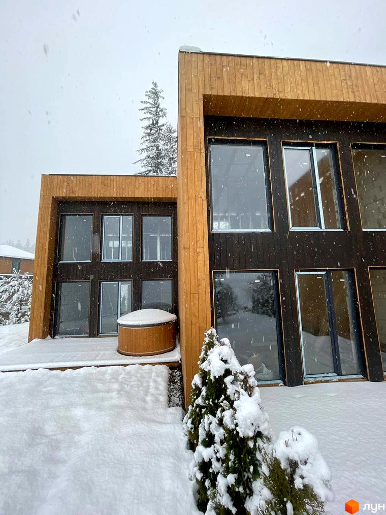 Котеджне містечко Агат Резорт & SPA. Двоповерховий будинок з комбінованим дерев'яним фасадом та великими вікнами, оточений снігом з дерев'яним джакузі на терасі.