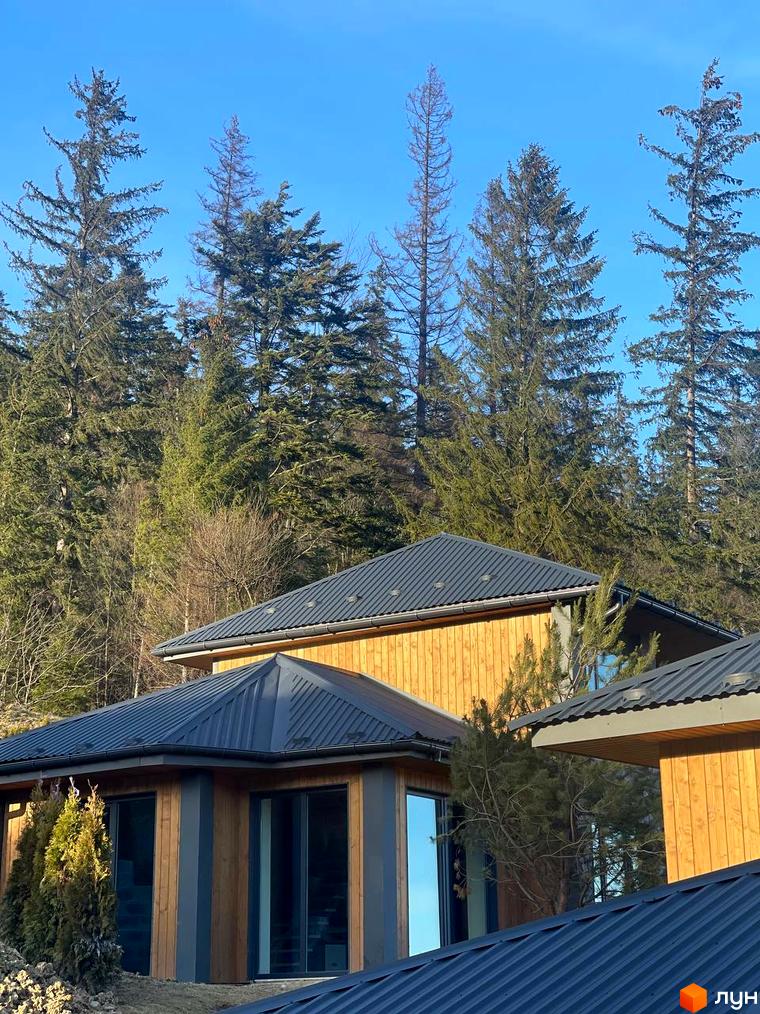 Котеджне містечко КМ Агат Резорт & SPA з дерев'яними фасадами та темними дахами на тлі лісу. Будинки розташовані на схилі гори під чистим блакитним небом.