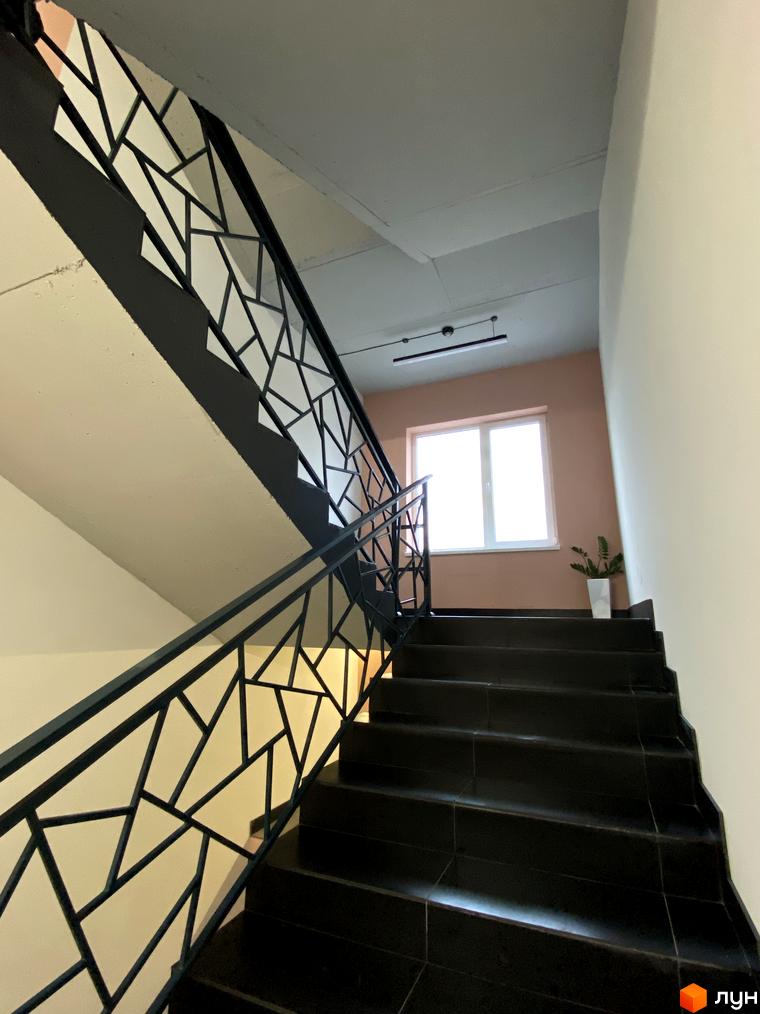 Сходи з чорними сходами та чорними поручнями з геометричним візерунком у клубному будинку Васильченка. На стіні біля вікна стоїть вазон із зеленою рослиною.