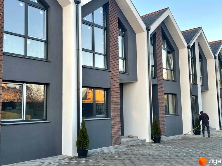 Зовнішній вигляд таунхаусів Dream Home з сірими та білими фасадами, цегляними вставками та темними вікнами. На прибудинковій території висаджені туї, а чоловік прибирає бруківку.