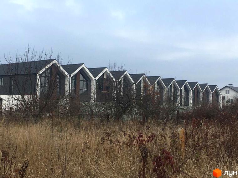 Ряд будинків Котеджна забудова Dream Home з темними дахами та світлими фронтонами, великими вікнами. Перед ними суха трава та чагарники.