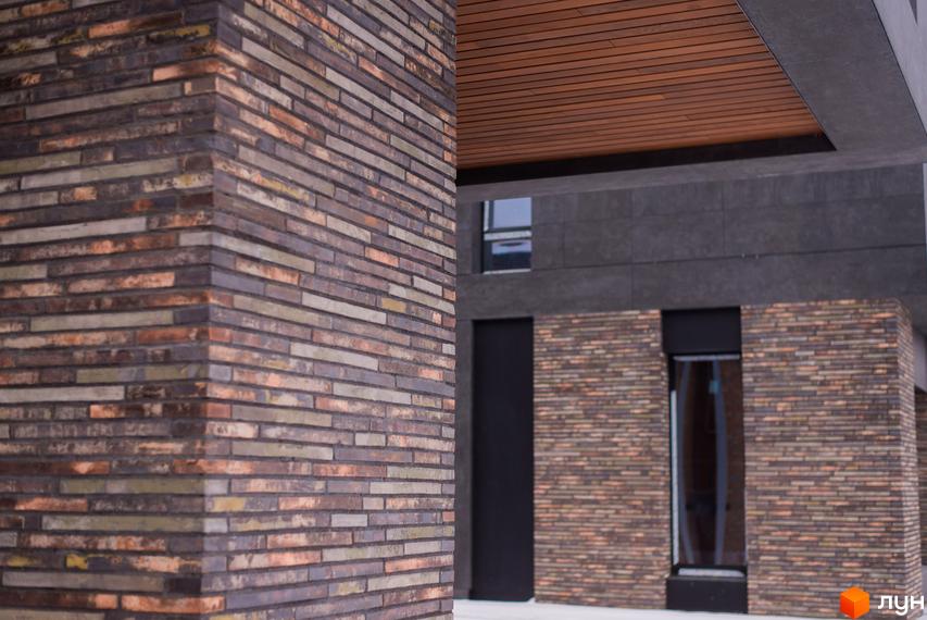Котеджне містечко КМ 9 ЕЛЕМЕНТІВ. Фасад будинку оздоблений цеглою різних відтінків та дерев'яними елементами під навісом. Біля входу розташовані скляні двері.