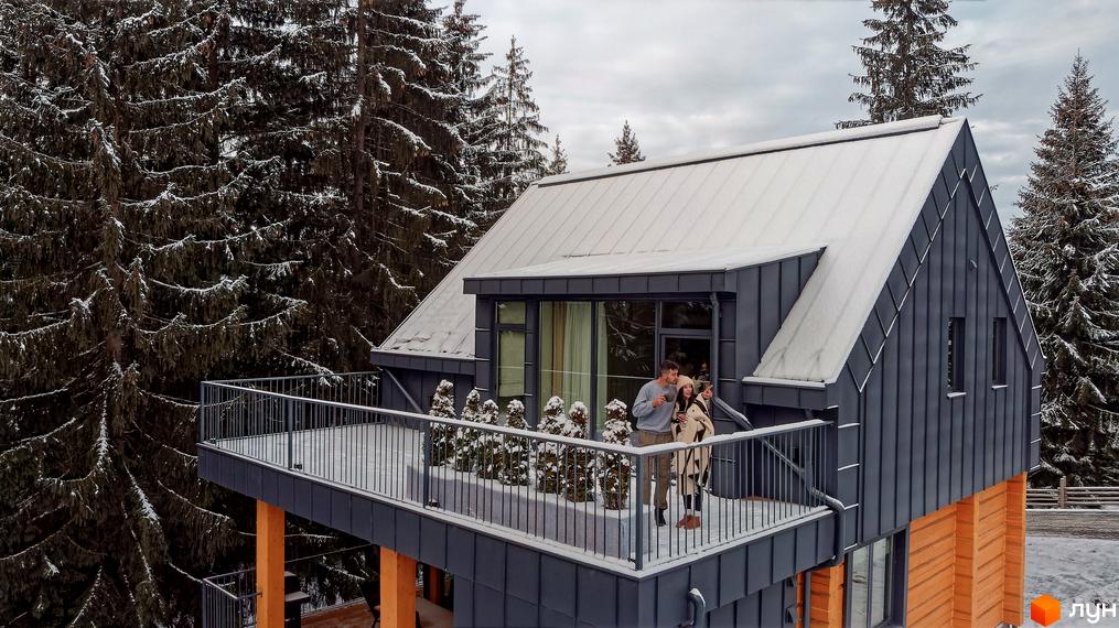 Вид на Котеджне містечко SKOGUR взимку: будинок з балконом, де відпочиває пара, навколо засніжені сосни.