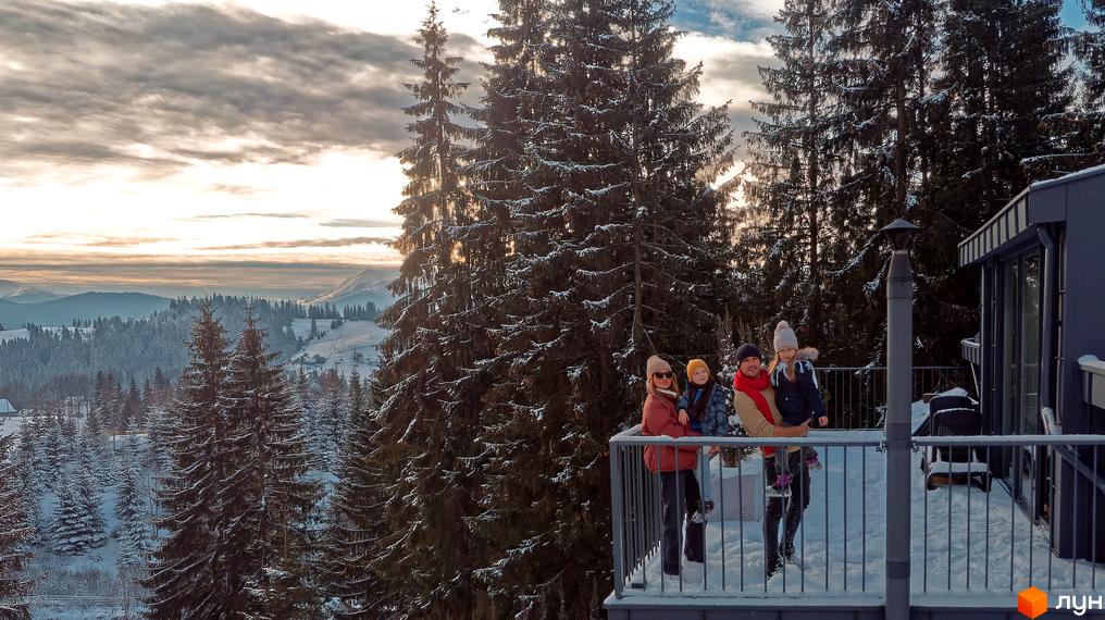 Родина на балконі Котеджного містечка SKOGUR на фоні засніженого лісу та гір. Зображення будинку та прибудинкової території.