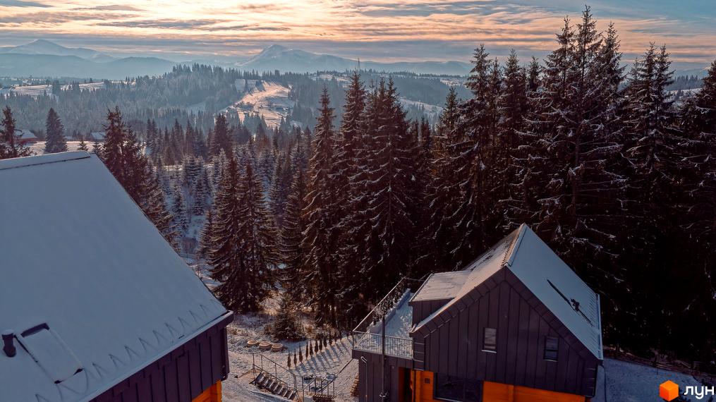 Зимовий пейзаж з Котеджного містечка SKOGUR. Будинки з темними дахами, вкриті снігом, на тлі засніжених гір та хвойного лісу.