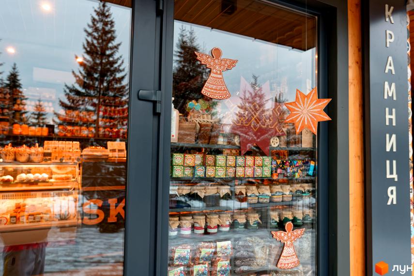 Вітрина крамниці з новорічним декором. На полицях видно різноманітні продукти та солодощі, а за вікном – зимовий ліс.