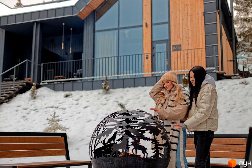 Зображення Котеджного містечка SKOGUR з будинком з великими вікнами та двома дівчатами біля декоративної сфери у сніжному дворі.