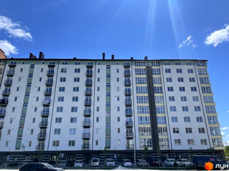 Зовнішній вигляд багатоповерхового будинку ЖК Квартал Гімназійний з білою фасадною частиною та зеленими елементами. Перед будинком розташована прибудинкова територія з автомобілями та деревами.