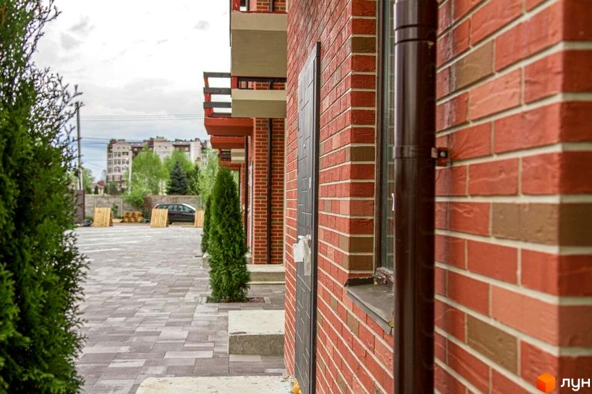 Цегляний будинок з балконами та темними дверима. На передньому плані доріжка з бруківки та зелені насадження.