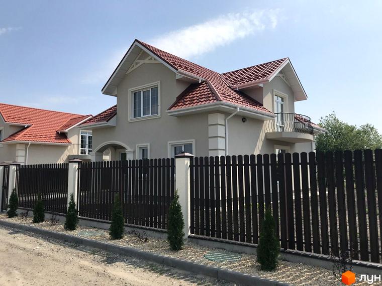 Котеджне містечко Зарічний Гай. Фасад будинку світло-бежевого кольору з червоним черепичним дахом, огороджений темним парканом. Поруч висаджені молоді ялинки.