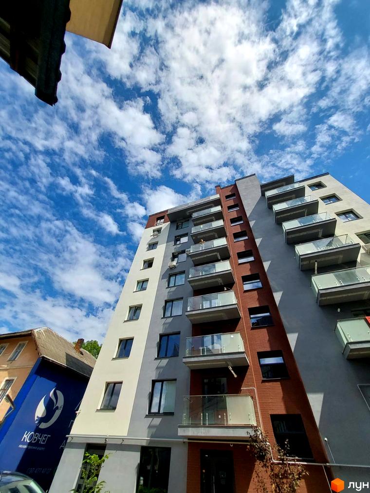 Клубний будинок Kovcheg Residence з цегляним та сірим фасадом, скляними балконами на тлі блакитного неба з хмарами. Поруч розташована будівля з синьою рекламою.