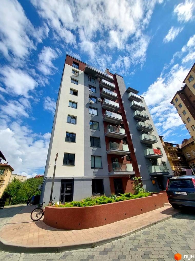 Клубний будинок Kovcheg Residence з сірими та білими фасадами та скляними балконами на фоні блакитного неба з хмарами. Поруч розташована прибудинкова територія з бруківкою та зеленими насадженнями.