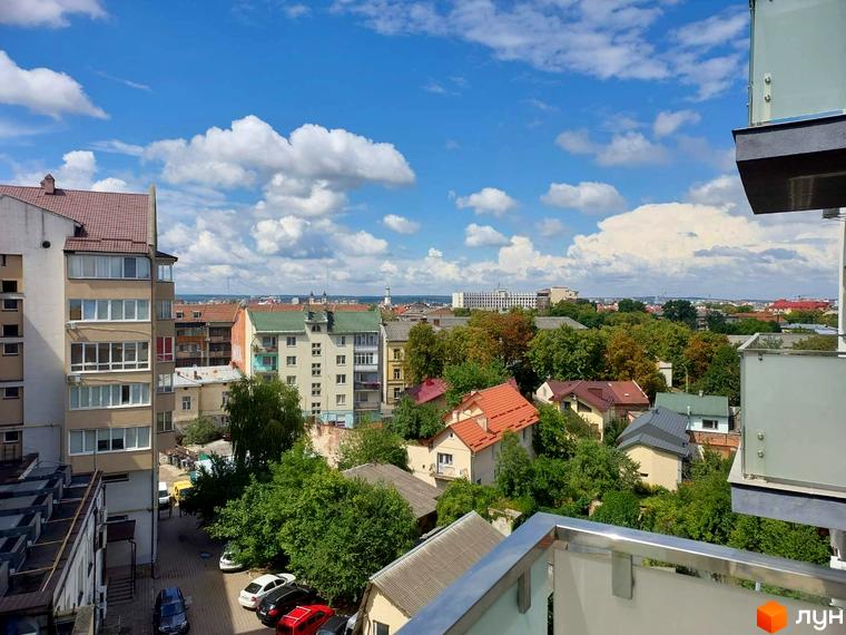 Вид на клубний будинок Kovcheg Residence з вікна, що виходить на міський пейзаж з багатоповерховими та приватними будинками, оточеними зеленню. На передньому плані видно балкон.