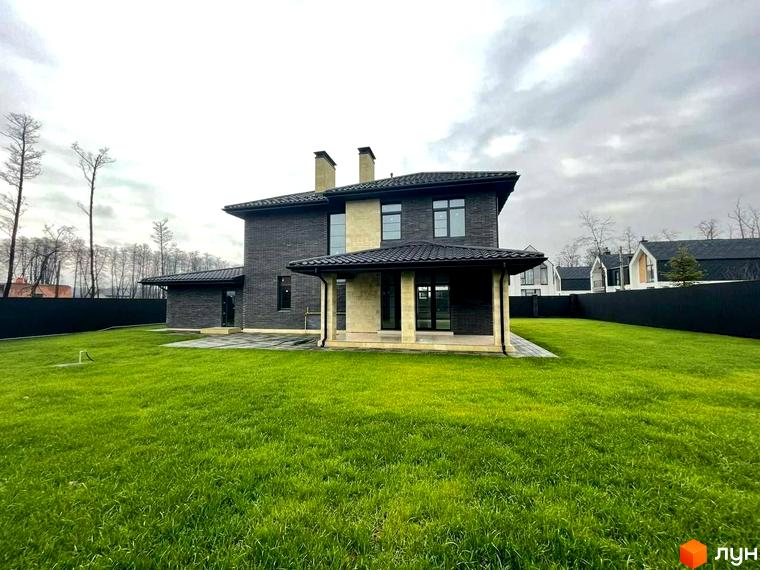 Котеджне містечко Spacious HOME з темно-сірою цегляною фасадною частиною, світлими кам'яними вставками та доглянутим зеленим газоном. Навколо будинку розташована огороджена територія з деревами та іншими будинками.