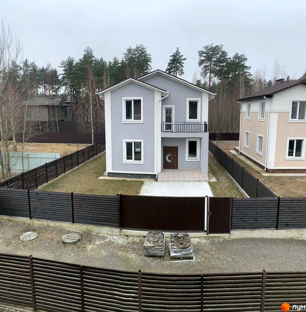 Зовнішній вигляд двоповерхового будинку в КМ ForestK сірого кольору з білими вікнами та балконом. Навколо будинку розташована темно-коричнева дерев'яна огорожа та зелений газон.