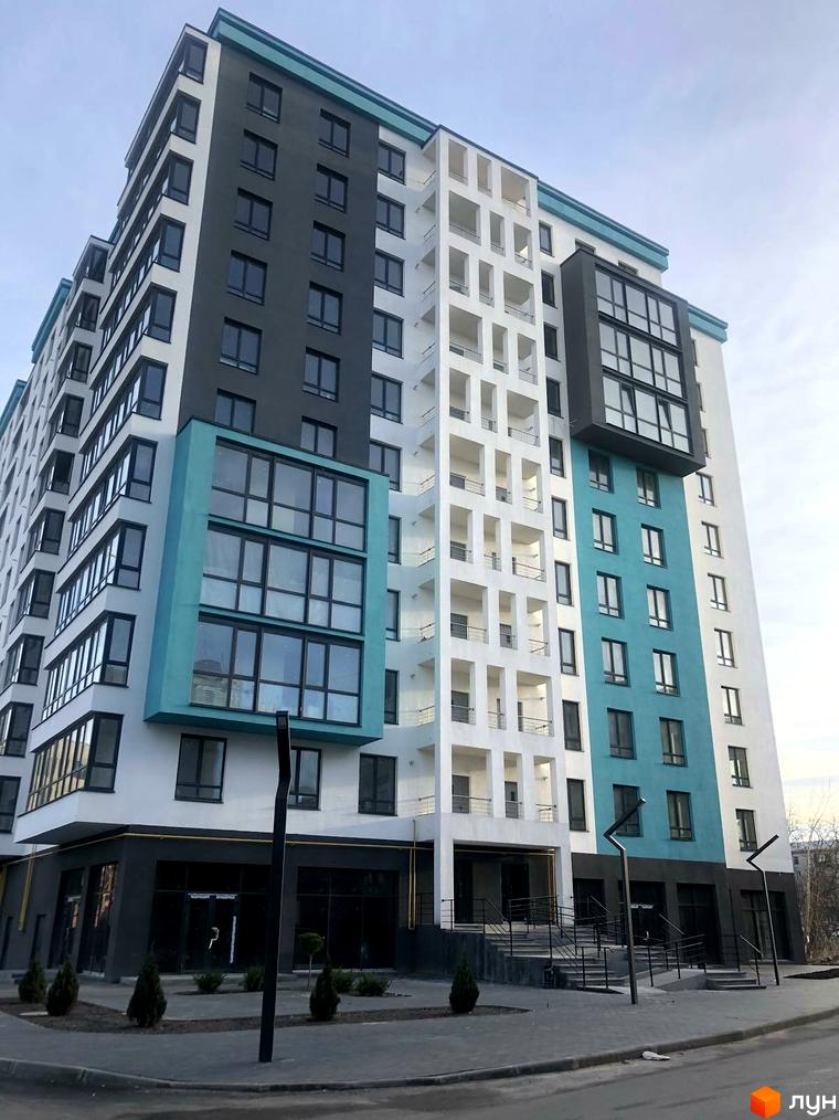 Комплекс SkyCity з білими та сірими фасадами, блакитними акцентами та великими вікнами. Перед будинком розташована прибудинкова територія з доріжками та зеленими насадженнями.