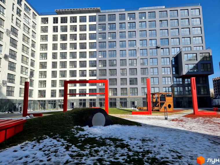 Багатоповерховий будинок Житлового комплексу Вільне Місто з фасадами світлого та темно-сірого кольору. На прибудинковій території видно дитячий майданчик, зелені пагорби та сніг.