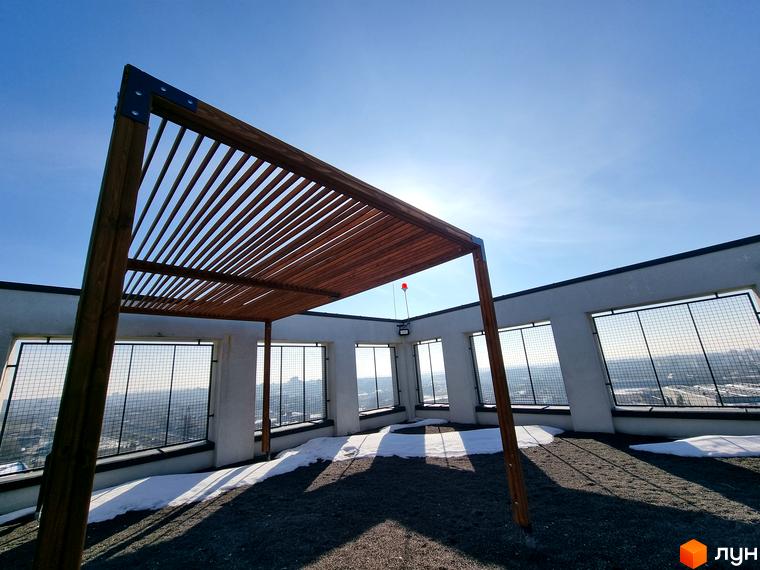 Тераса на даху Житлового комплексу Вільне Місто з дерев'яною перголою, гравійним покриттям та панорамним видом на місто через вікна з решітками.