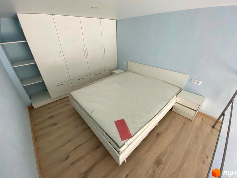 Спальня з білим ліжком та шафою у ЖК Сузір'я 2020. На стіні блакитні шпалери, на підлозі дерев'яне покриття.