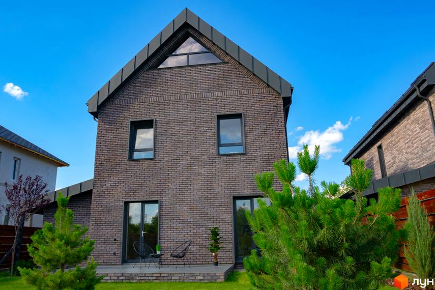 Котеджне містечко КМ BritanHouse. Зовнішній вигляд двоповерхового будинку з цегляним фасадом та зеленою прибудинковою територією під блакитним небом.