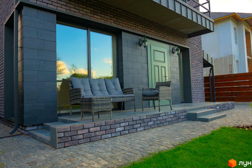 Зображення будинку в Котеджному містечку BritanHouse з терасою, меблями та доріжкою з бруківки. Навколо будинку газон та зелені насадження.