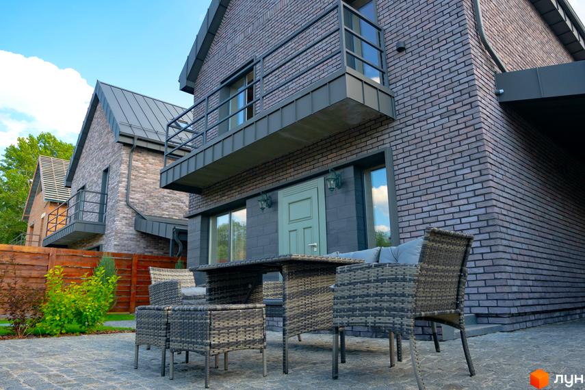 Цегляний будинок з балконом та терасою, обладнаною плетеними меблями. На подвір'ї викладена бруківка, видно зелені насадження.