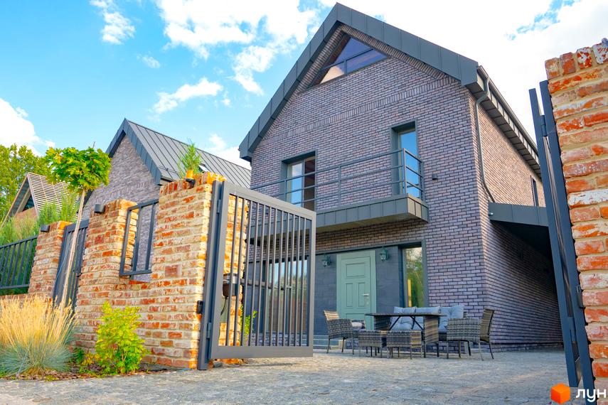 Котеджне містечко BritanHouse. Фасад будинку з цегли, металевий дах, балкон, вхідна група з дверима та вікнами. Прибудинкова територія з цегляним парканом, воротами, зоною відпочинку та озелененням.