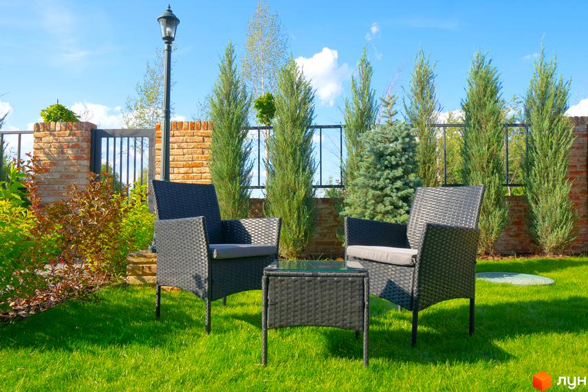 КМ BritanHouse: затишна зона відпочинку з плетеними меблями на доглянутому газоні біля цегляного паркану та зелених насаджень.