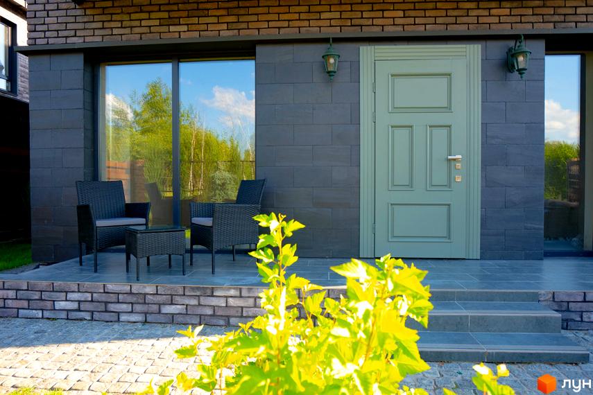 Зовнішній вигляд будинку КМ BritanHouse з сірою плиткою та зеленою дверима. На прибудинковій території розташовані крісла та столик.