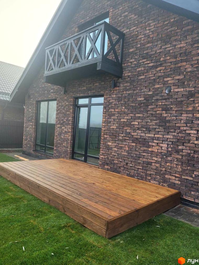 Котеджне містечко Панська Баварія: цегляний будинок з дерев'яною терасою та зеленим газоном. Зовнішній вигляд новобудови та прибудинкової території.