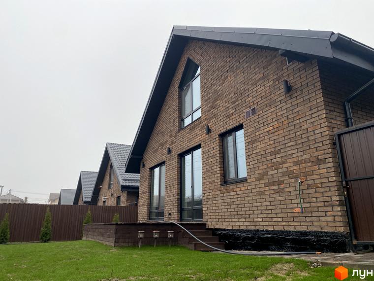 Котеджне містечко Панська Баварія. Фасад будинку з коричневої цегли, вікна, дах та паркан виконані у темних тонах. Зелений газон та дерева біля будинку.